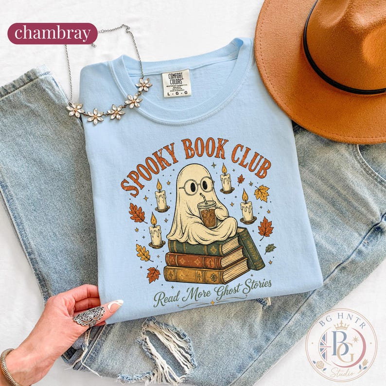Pode incluir: Uma t-shirt azul-clara com um gr&aacute;fico de um fantasma de &oacute;culos, sentado numa pilha de livros enquanto segura uma bebida. O texto diz SPOOKY BOOK CLUB e Read More Ghost Stories, rodeado por velas e folhas de outono. Um colar de flores prateado e um chap&eacute;u bege s&atilde;o vis&iacute;veis.