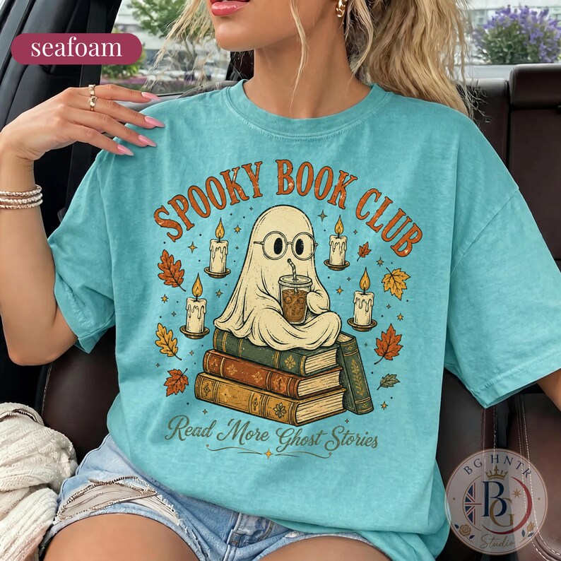 Pode incluir: Uma t-shirt verde espuma do mar com um gr&aacute;fico de um fantasma de &oacute;culos, segurando um copo de caf&eacute; e sentado numa pilha de livros. O texto "SPOOKY BOOK CLUB" e "Read More Ghost Stories" &eacute; rodeado por velas acesas e folhas de outono.