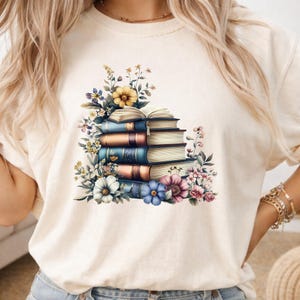 May include: A cream-colored t-shirt featuring a graphic print of a stack of five books with an open book on top. The stack is surrounded by various colorful flowers including yellow, white, blue, and pink blossoms. The design is centered on the chest.