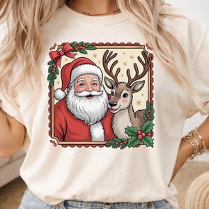 May include: A cream-colored t-shirt featuring a square graphic of Santa Claus with a white beard and red hat standing next to a brown reindeer. The illustration is framed by a red scalloped border with green holly leaves and red berries.