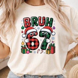 May include: A cream-colored t-shirt featuring a graphic with the text BRUH and the numbers 67 in red and green buffalo plaid. The numbers have cartoon eyes and wear Santa hats, surrounded by colorful Christmas ornaments, gifts, and snowflakes.