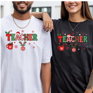 May include: A white t-shirt and a black t-shirt, each featuring a graphic print of the word TEACHER in festive patterns. The design includes a red mug with cinnamon sticks, a reindeer ornament, and scattered snowflakes in red and green colors.