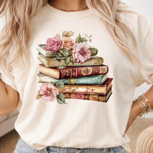 May include: A cream-colored t-shirt featuring a graphic print of a stack of five vintage-style books adorned with various pink and peach-colored roses and green leaves. The books have detailed, ornate spines and aged pages, creating a layered composition.