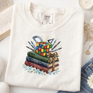 May include: A cream-colored t-shirt featuring a graphic of a colorful Rubik's cube resting on a stack of three books titled Advanced Calculus, Linear Algebra, and Mathematics & Logic, accented with a leopard print bow and blue flowers.