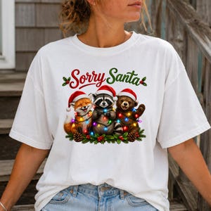 May include: A white t-shirt featuring a graphic of a fox, a raccoon, and a bear cub wearing red Santa hats. The animals are wrapped in colorful Christmas lights and surrounded by pine branches and holly. The text Sorry Santa is printed above.