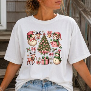 May include: A white short-sleeve t-shirt featuring a central graphic print of Christmas-themed illustrations in shades of pink and green, including Santa Claus, a decorated pine tree, a reindeer, a snowman, a gift box, bells, and bows.
