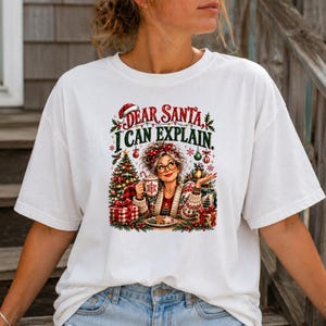 May include: A white short-sleeved t-shirt featuring a graphic print of a person with glasses wearing a patterned cardigan, holding a mug, and sitting by a Christmas tree. The text reads Dear Santa I Can Explain in red and green lettering.