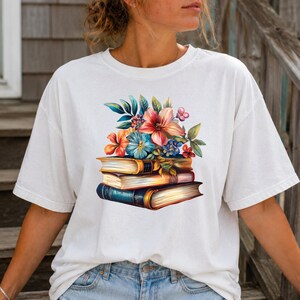 May include: A white short-sleeve t-shirt featuring a graphic print of a stack of three books topped with a vibrant arrangement of colorful flowers including pink, orange, and blue blossoms with green leaves, centered on the chest area.