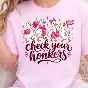 May include: A light pink t-shirt featuring a graphic of four white ducks. The ducks wear pink boxing gloves, scarves, and bows, with one holding a pink flag that says HOPE. The text below the ducks reads check your honkers in dark pink script.