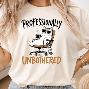 May include: A cream-colored t-shirt featuring a graphic of a cat wearing sunglasses, sitting in an office chair, and holding a steaming mug. The text above reads PROFESSIONALLY and the text below reads UNBOTHERED in bold, dark lettering.