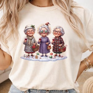 May include: A cream-colored t-shirt featuring a graphic of three elderly individuals with white hair, wearing winter clothing and glasses, each holding a wine glass. They stand on a snowy base decorated with ornaments, a gingerbread man, and candy canes.