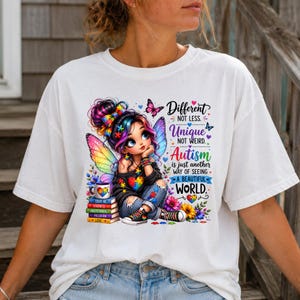 May include: A white t-shirt featuring a colorful graphic of a character with butterfly wings, sitting on a stack of books. The text reads: Different NOT LESS. Unique NOT WEIRD. Autism is just another way of seeing A BEAUTIFUL WORLD.