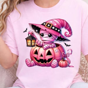 May include: A light pink t-shirt featuring a graphic of a mummy wearing a pink witch hat, sitting inside a large pink jack-o'-lantern. The mummy holds a small black lantern, and the design includes small black bats and a smaller pink pumpkin.