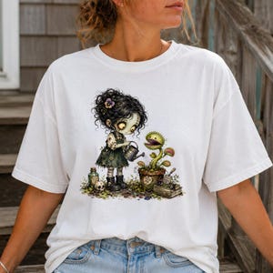 May include: A white short-sleeve t-shirt featuring a graphic illustration of a dark-haired character in a green dress watering a potted Venus flytrap. The pot is labeled with the text FEED ME. Additional details include a small skull, a glass bottle, and a trowel.