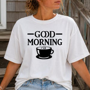May include: A white short-sleeve t-shirt featuring black text that reads GOOD MORNING with a horizontal line on each side of the word GOOD. Below the text is a black graphic of a coffee cup with a small heart shape inside the foam.