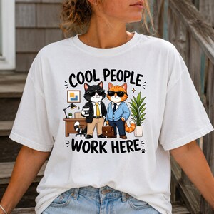 May include: A white t-shirt featuring a graphic of two cats wearing sunglasses and business attire. One cat is black and white, the other is orange. They stand in an office setting with a desk, lamp, and plant. The text reads COOL PEOPLE WORK HERE.