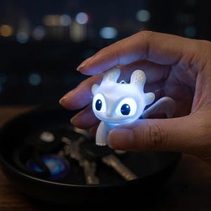 May include: A small, white, glowing dragon-shaped keychain charm with large, expressive eyes and textured ears. The figure emits a bright internal light, highlighting its smooth surface and rounded features, designed to attach to a set of metal keys.