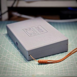 May include: A rectangular gray plastic case with a textured surface featuring an embossed graphic of a handheld gaming console and the text THOR. A thin orange cord with a matching orange plastic toggle is attached to the side of the case.