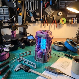 May include: A purple 3D printed sci-fi containment unit with a glowing blue spiral core, featuring the text MYC8-085-YC. The object sits on a green cutting mat alongside a digital caliper, screwdrivers, filament spools, and a 3D printer.