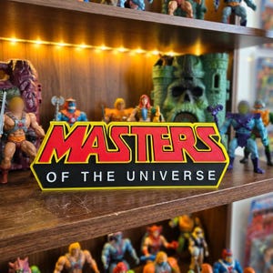 Può includere: Un'insegna con il logo Masters of the Universe caratterizzata da scritte rosse in grassetto con un bordo giallo su sfondo nero. L'insegna è posizionata su uno scaffale di legno davanti a varie action figure, inclusa una struttura a castello verde.