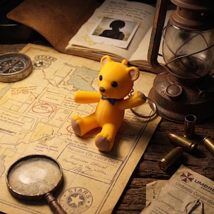 May include: A yellow bear keychain with a small blue bowtie, black eyes, and light brown paw pads. The bear is positioned on a map of Raccoon City Police Dept, surrounded by a compass, a magnifying glass, a lantern, and scattered brass bullet casings.