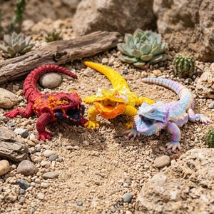 Peut inclure: Trois figurines de lézards articulées avec des écailles texturées. L'une est rouge et noire, l'une est jaune vif et orange, et l'une présente un dégradé arc-en-ciel pastel. Chaque lézard a la gueule ouverte, quatre pattes et une longue queue.