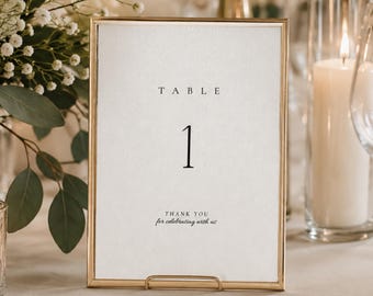 Pode incluir: Um sinal de número de mesa retangular vertical numa moldura fina de metal dourado. O cartão branco apresenta a palavra TABLE em fonte serifada preta acima do número grande 1, com o texto THANK YOU for celebrating with us na parte inferior.