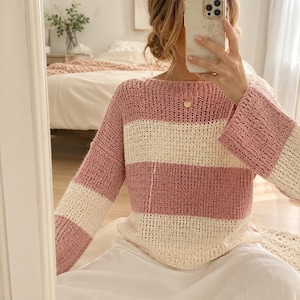 May include: A long-sleeved, loose-fitting sweater featuring a horizontal striped pattern in alternating dusty rose pink and cream white. The garment is crafted with a visible open-knit crochet texture, a wide boat neckline, and straight sleeves.