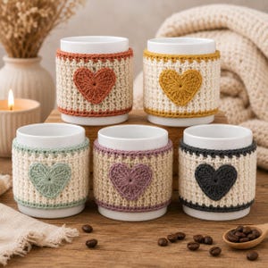May include: Five white ceramic mugs, each wrapped in a textured, cream-colored crochet cozy. Each cozy features a single, solid-colored heart centered on the front in shades of rust orange, mustard yellow, sage green, dusty purple, and charcoal gray.