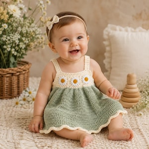 May include: A crochet dress featuring a sage green skirt and a cream-colored bodice adorned with three white and yellow daisy motifs. The dress has thin straps and a scalloped hem. A matching cream-colored headband with a small bow is included.