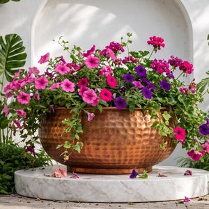 Pode incluir: Uma grande taça de cobre martelado, redonda, cheia de flores de petúnia vibrantes em tons de rosa, magenta e roxo, com trepadeiras de hera verde, assente numa base circular de mármore branco contra uma parede branca com um nicho.