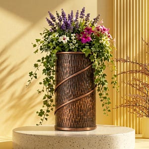 May include: A cylindrical, hammered copper-toned metal planter featuring a spiraling raised band design. The planter is filled with an arrangement of purple lavender, pink and white petunias, and trailing green ivy foliage.