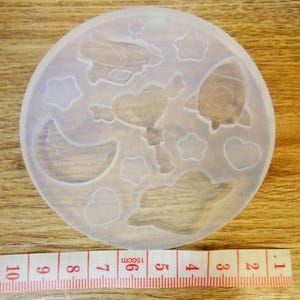 Op de afbeelding: Een heldere, ronde siliconen mal met verschillende ruimtethema-vormen, waaronder een raket, vliegtuig, sterren, harten en een halve maan. Een meetlint met centimeter markeringen is zichtbaar eronder.