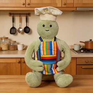 May include: A plush toy shaped like a round, light green creature wearing a white chef hat and a colourful apron with vertical stripes in red, orange, yellow, blue, and white, featuring a front pocket with horizontal stripes in the same colours.