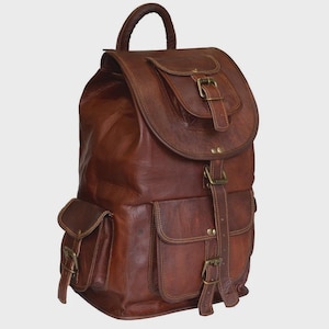 Peut inclure: Un sac à dos en cuir marron doté d'une poignée supérieure, d'un rabat principal avec fermeture à boucle et de trois poches extérieures avec sangles à boucle. Le sac présente des coutures visibles et des ferrures métalliques structurées.