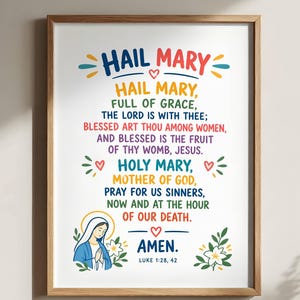 May include: A framed wall art print featuring the Hail Mary prayer in colourful text. The design includes a small illustration of Mary with a yellow halo, green leaves, and white flowers at the bottom, with the text Luke 1:28, 42 at the base.