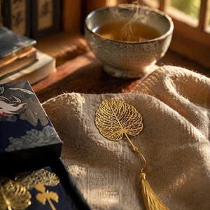 Peut inclure: Un marque-page en métal doré en forme de feuille squelettée avec un motif de nervures en filigrane délicat. Le marque-page présente une fine tige métallique attachée à un long pompon en soie assorti de couleur dorée sur un tissu texturé.