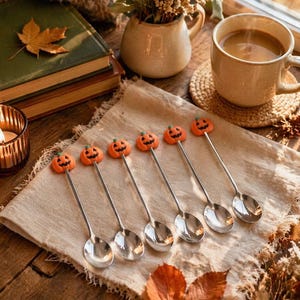 Peut inclure: Un ensemble de six cuillères en métal argenté, chacune dotée d'une petite breloque citrouille orange sculptée avec un visage noir gravé, fixée au sommet du manche. Les cuillères sont disposées en rangée sur une serviette en tissu texturé.