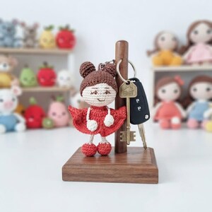 May include: A crochet doll keyring with brown hair in two buns, wearing a red dress and matching shoes, hanging on a wooden stand alongside a metal house-shaped key tag and a black car key fob. The stand has a square base and a vertical post.