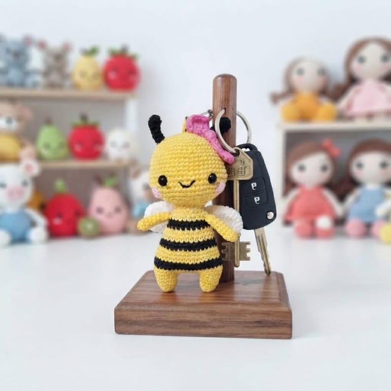 May include: A yellow and black crocheted bee keyring with a small pink flower accessory, attached to a metal ring and a black car key fob, displayed on a vertical wooden stand with a square base, featuring a soft, textured yarn construction.
