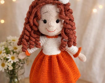May include: A handmade crochet doll featuring curly auburn yarn hair with a white bow, a cream-coloured top, and a vibrant orange skirt. The doll has embroidered facial features, white ruffled trim at the ankles, and brown shoes, standing upright.