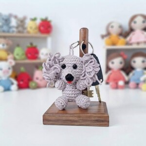 May include: A light grey crocheted dog keyring with curly ears, black eyes, and a small red tongue, hanging from a metal ring on a vertical wooden post attached to a square wooden base. The dog is crafted with visible yarn stitches.
