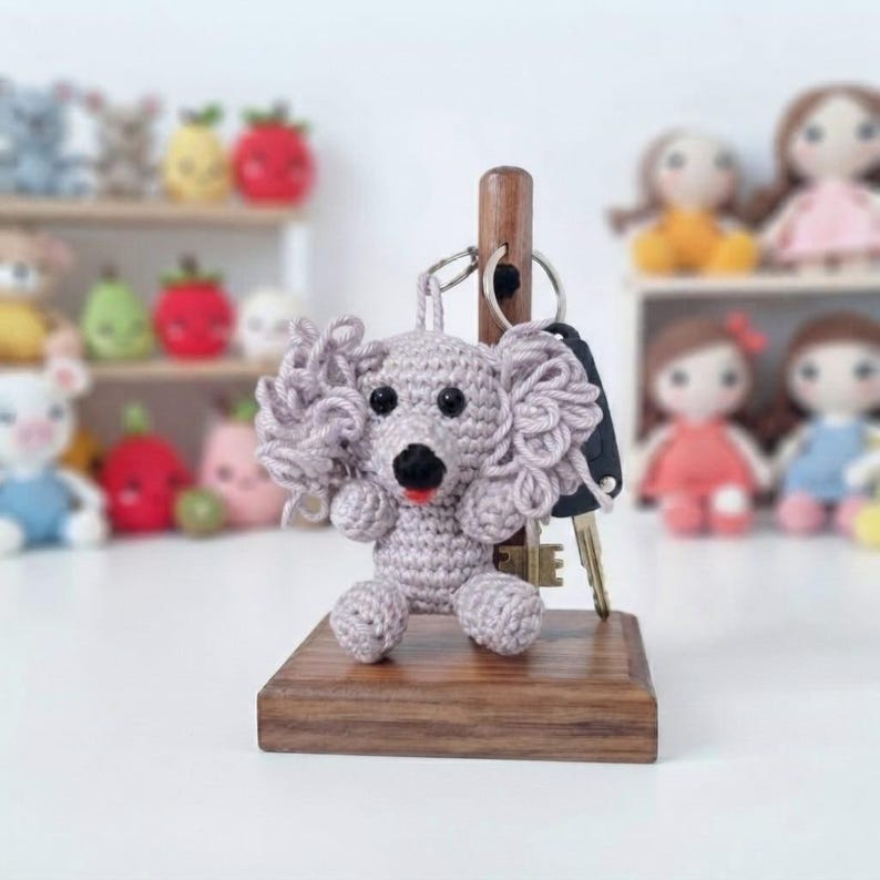 May include: A light grey crocheted dog keyring with curly ears, black eyes, and a small red tongue, hanging from a metal ring on a vertical wooden post attached to a square wooden base. The dog is crafted with visible yarn stitches.