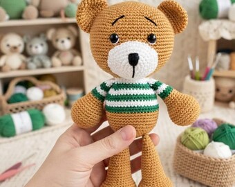 May include: A crocheted teddy bear toy with light brown yarn, a white muzzle, and a black embroidered nose. The bear wears a green and white striped sweater. It features round ears, black plastic eyes, and thin, dangling arms and legs.