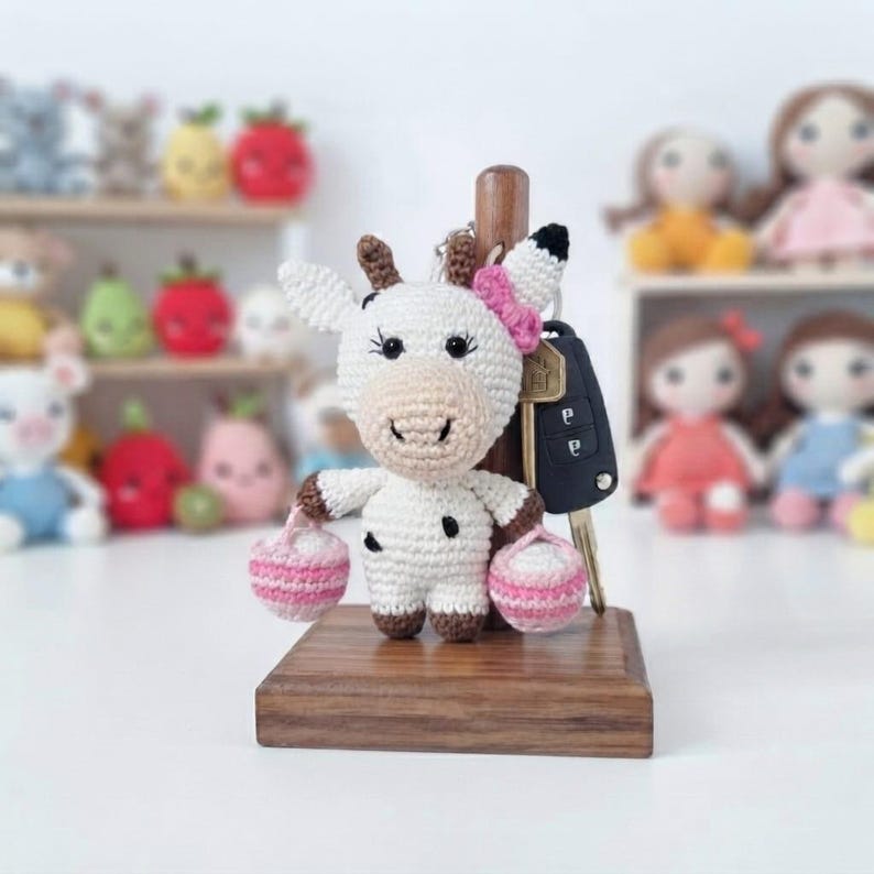 May include: A white and brown crocheted cow keyring featuring a small pink bow on its head and two tiny pink and white striped baskets held in its hands. The cow is attached to a black car key fob and stands on a rectangular wooden base.