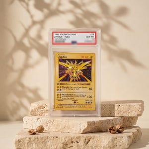 May include: A 1999 Pokemon Game Zapdos Holo trading card graded GEM MT 10, encased in a clear protective plastic slab. The card features a yellow background, a holographic illustration of the electric bird, and text including 90 HP and attacks.