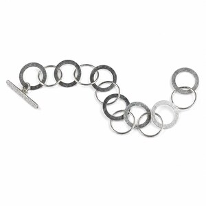 May include: A bracelet composed of alternating dark gray and silver-toned metal rings linked together in a wavy pattern. The piece features a toggle clasp with a rectangular bar and a circular ring, showcasing a textured, hammered finish throughout.