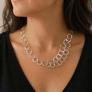 May include: A silver-toned necklace composed of numerous interconnected circular metal rings. The chain features a single row of rings that transitions into a wider, multi-layered cluster of overlapping circles on one side of the collarbone.