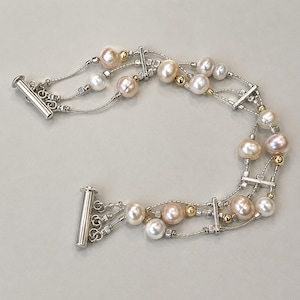 May include: A multi-strand bracelet featuring white and soft pink freshwater pearls interspersed with small gold-tone beads and silver-tone metal spacers. The design includes three delicate silver-tone chains secured by a cylindrical toggle clasp.
