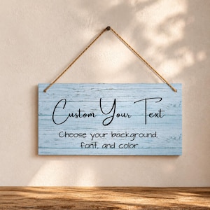 May include: A rectangular light blue wooden sign with a weathered texture, suspended by a brown rope. The sign features the black text "Custom Your Text" and "Choose your background, font, and colour." in a combination of script and sans-serif fonts.